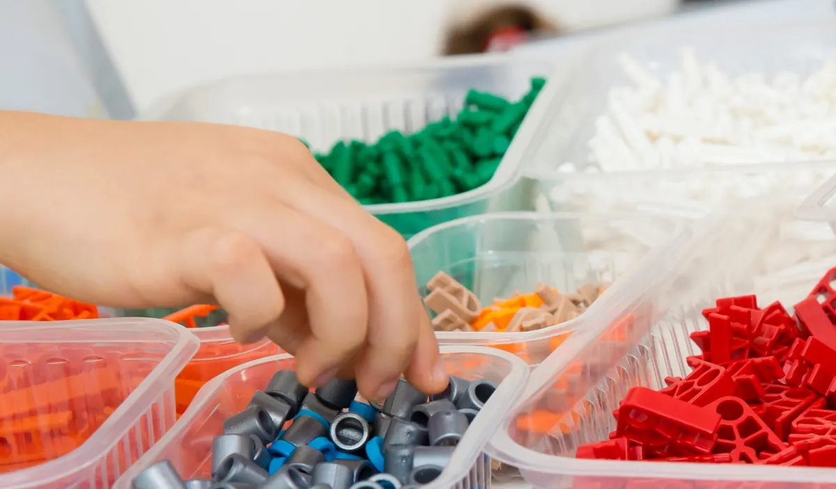 Mão de aluno pegando conectores K'NEX em bandejas separadas por cor